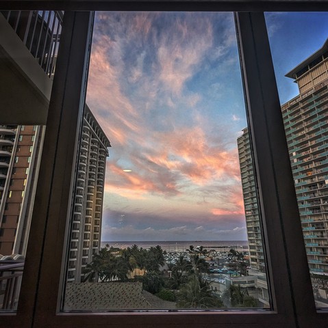 Looking through a window on the eighth floor seven minutes before sunrise the blue sky above the ocean is full of swirling pink clouds. Tall hotels flank the left and right, and a cluster of palm trees is in the middle.