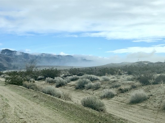 A wide shot of a desert landscape from the side of the road with sparse vegetation, sand, & mountains partially shrouded in mist under a blue sky with clouds.