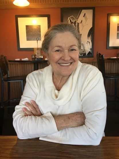 Upper body & head photo of a pretty woman in her mid-70s. She’s sitting at a restaurant table. She’s smiling, has her hair pulled back, & is wearing a cream colored cowl neck sweater.