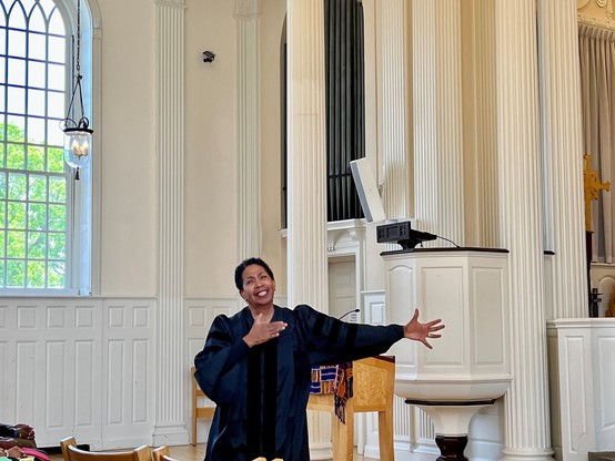 A Black woman with an outstretched arm, beaming a smile, wearing a dark blue and black liturgical robe, in front of a pulpit in a chapel.