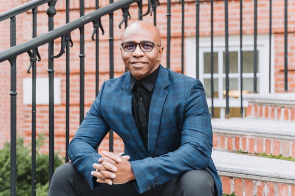 A Black man with a bald head, wearing black rimmed glasses, and a checked blue sport coat and black dress shirt, sitting on steps in front of a brick building and wrought-iron railing.