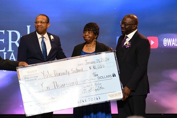 Three people, two men and a woman, stand together holding an oversized bank check made out to Yale Divinity School for ten-thousand dollars. The check is handed to them by a person off frame to the left.