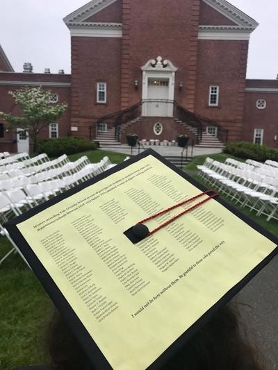 In the foreground, a graduate mortar board hat with a yellow paper attached to the top. The paper has columns of names printed on it. In the background, a red brick chapel exterior and white folding chairs set up for graduation.