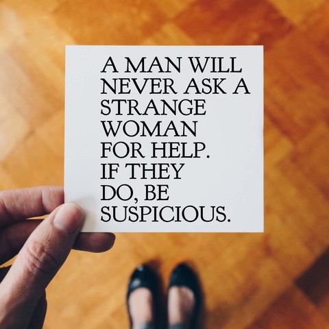 A hand is holding a card with text that reads A MAN WILL NEVER ASK A STRANGE WOMAN FOR HELP. IF THEY DO, BE SUSPICIOUS.