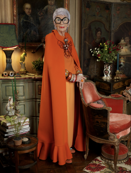 Amazing professional full-body portrait of a beautiful old woman with short grey hair & huge black rimmed round glasses. She's wearing a FABULOUS orange cape with tons of amazing jewelry, & she standing with her hands on a velvet armchair in a room stuffed full of gorgeous things.