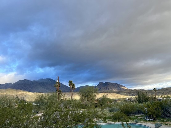 Wide photo taken from our back patio of the dark mountains with heavy clouds, stormy skies & desert plants & palm trees in the foreground