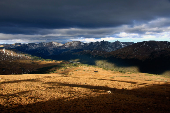 Landscape photo. A view of The Never Summer Range looking west from Trail Ridge Road in Rocky Mountain National Park, shortly after sunrise. The mountains across the middle of the frame are lightly covered with snow at the higher elevations, still in shadow below. The foreground is tundra grass lit by the sun, with shadows reaching across. The sky is dark grey above, with blue sky visible just above the mountains. At the extreme right middle of the image, if you zoom in you can see the faint ma…