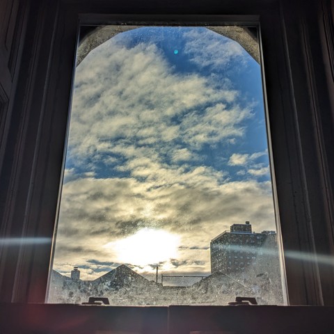 Looking through an arched window an hour after sunrise the sun is creating a dramatic curved lens flare across a blue sky full of mottled white and yellow clouds. Pointed roofs of Harlem brownstones are silhouetted across the street, and a taller apartment building can be seen in the distance. The window is showing a lot of backlit grime. 