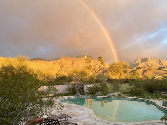 Photo taken from our back patio showing a massive rainbow in the cloudy grey sky with a faint 2nd rainbow, morning sunlit mountains in the background, & the reflection of the rainbow in our pool.