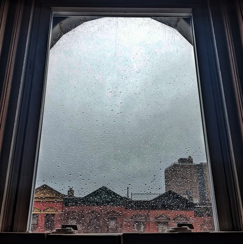 Looking through an arched window four hours after sunrise the sky is full of grey rain clouds and the glass is covered in rain drops. Pointed roofs of Harlem brownstones with red brickwork are across the street, and a taller apartment building can be seen in the distance. 