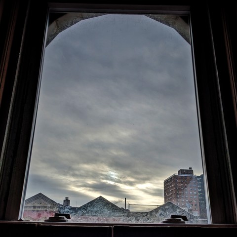 Looking through an arched window an hour after sunrise the sky is full of churning grey clouds and the sun is breaking through in the lower sky. Pointed roofs of Harlem brownstones with red brickwork are silhouetted across the street, and a taller apartment building can be seen in the distance. The window is grimy on the bottom.