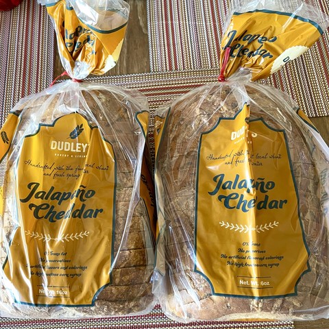 Two loaves of fresh Dudley’s Jalapeno Cheddar bread in transparent & yellow packaging on a striped placemat