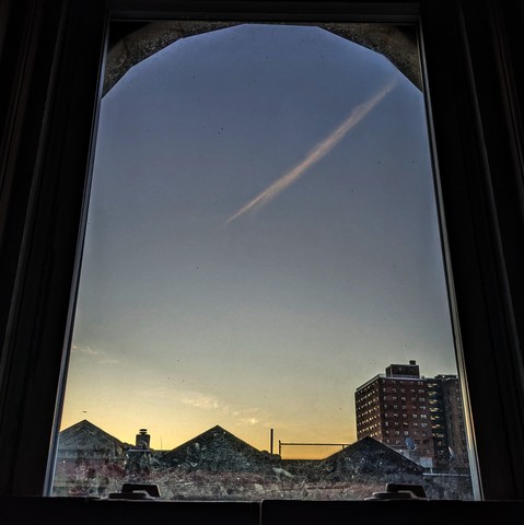 Looking through an arched window at the moment of sunrise the sky fades from parchment at the horizon to deep blue above. A jet trail makes a diagonal streak down from the top right. Pointed roofs of Harlem brownstones with red brickwork are silhouetted across the street, and a taller apartment building can be seen in the distance. The window is grimy on the bottom. 