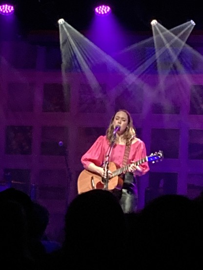 Sarah Jarosz is front and center, wearing a pink shirt and black leather pants. She’s playing an acoustic guitar and singing into a microphone onstage with stage lights casting purple and green beams in the background.