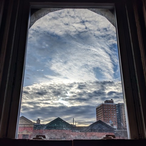 Looking through an arched window an hour after sunrise the blue sky is full of complex swirling white and grey clouds. Pointed roofs of Harlem brownstones with red brickwork are  across the street, and a taller apartment building can be seen in the distance. The window is grimy on the bottom. 