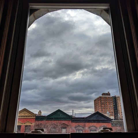 Looking through an arched window thirty eight minutes after sunrise the sky is full of complex grey clouds. Pointed roofs of Harlem brownstones with red brickwork are across the street, and a taller apartment building can be seen in the distance.