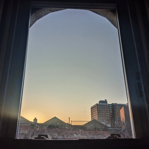 Looking through an arched window fifteen minutes after sunrise the cloudless sky fades from peach at the horizon to robin-egg blue up above. Pointed roofs of Harlem brownstones  are silhouetted across the street, and a taller apartment building can be seen in the distance. The window is grimy on the bottom. 