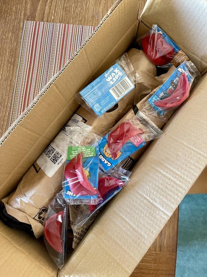 Photo of open cardboard box sitting on our dining room table, it's holding a brown paper wrapped tripod & a bunch of individually packaged Wacko-Wax red wax lips