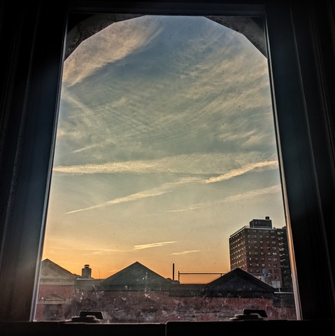 Looking through an arched window six minutes after sunrise the sky fades from peach at the horizon to slate blue up above and is criss-crossed by wispy white clouds. Pointed roofs of Harlem brownstones with red brickwork are silhouetted across the street, and a taller apartment building can be seen in the distance. The window is grimy on the bottom.