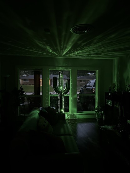Photo of a dimly lit room with a giant metal saguaro cactus sculpture partially lit with green LED lights displaying patterns on the ceiling in front of a window. A sofa is in the foreground and there’s a view of the outdoors through the window.
