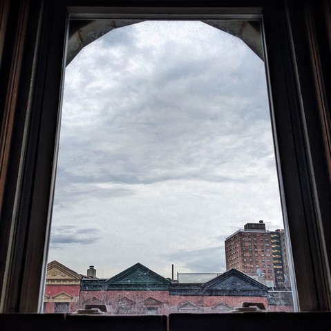 Looking through an arched window thirteen minutes after sunrise the sky is full of indistinct light gray clouds. Pointed roofs of Harlem brownstones with red brickwork are across the street, and a taller apartment building can be seen in the distance.