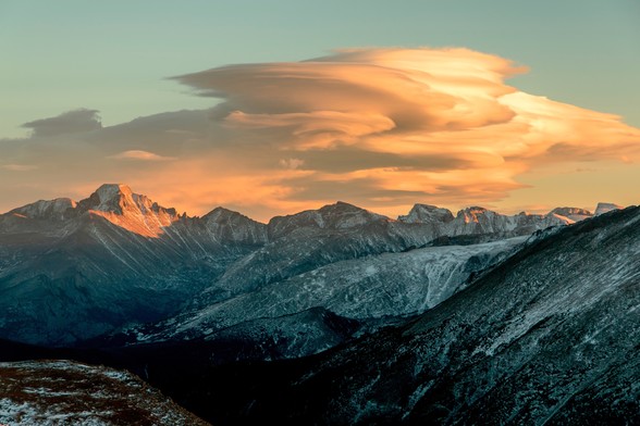 Panoramic photo. Longs Peak at sunset, viewed from Trail Ridge Road in Rocky Mountain National Park. A range of mountains stretches across the frame, a light covering of snow helps them to show in the growing shadows, as Longs Peak glows orange in the last of the sunlight. Billowing lenticular clouds rise up above the mountains, glowing in the orange light as well. October 2014.