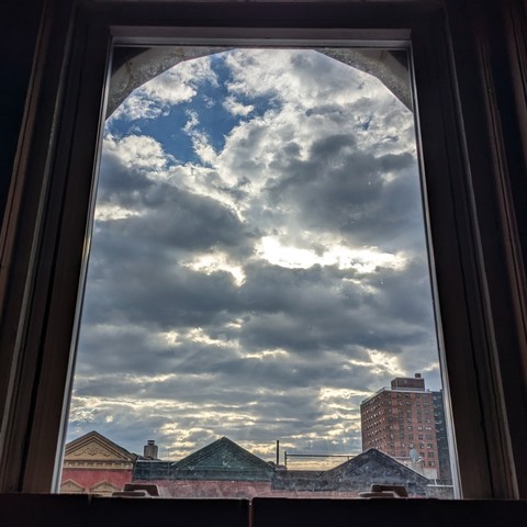 Looking through an arched window two hours after sunrise the sky is full of chunky gray clouds and a patch of blue sky is peeking through. Pointed roofs of Harlem brownstones with red brickwork are across the street, and a taller apartment building can be seen in the distance.