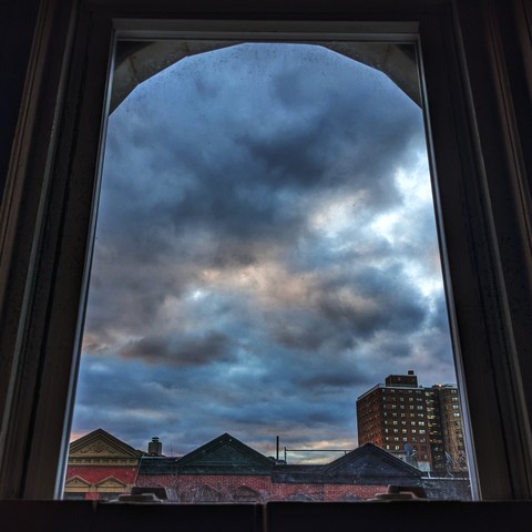 Looking through an arched window eleven minutes after sunrise the blue sky is full of complex gray and white clouds. Pointed roofs of Harlem brownstones with red brickwork are  across the street, and a taller apartment building can be seen in the distance.