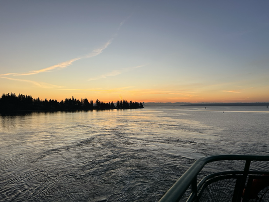 The sunrise glows orange and yellow on the horizon with blue skies an above and the waters of Eagle Harbor below. Trees in the shore are silhouetted and the railing of a Washington state ferry is in the foreground. 