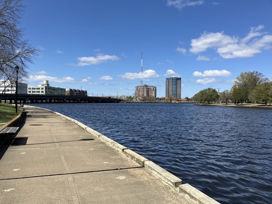 A riverside walkway with a view of a city skyline and a clear blue sky with a few clouds.