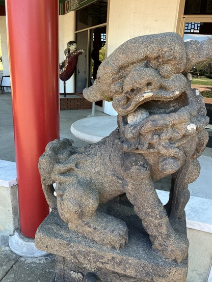 A worn stone lion sculpture guards the front of an unseen pagoda, a red pillar in view. 