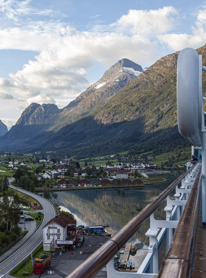 We're on the top deck of a cruise ship that's docked in a port at one end of a fjord. We can see a little of the fjord water in the direction along the side of the ship we're looking before it reaches a flat valley of land between a sharply rising mountainous range that heads further into the distance, carrying on the line of fjord mountains that must line the water behind us. There's a feel of summer about the scene with a blue sky and white, puffy clouds but there's also a little bit of a chill as most of the fjord valley including our position on the ship is in shade. We can see shadows of an unseen range of hills and mountains out of view on the left being cast up the sides of the mountains on the right so the sun must be quite low in the sky to the left of view. This is early in the morning. The flat bottom of the the valley is dotted with houses and shops, all only a couple of storeys high and with most of them close to the water to form a small town or village. A winding road passes by the ship on the left and we can see between it and our vessel there's a three storey high guest house and tables and parasols out for an attached bar.