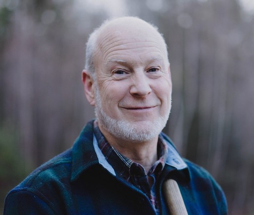 A man with a white beard and bald head smiles at the camera in an outdoors setting.