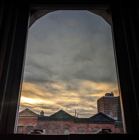 Looking through an arched window twenty-two minutes after sunrise the sky is full of churning blue-gray clouds as the sun is breaking through on the lower left. Pointed roofs of Harlem brownstones with red brickwork are across the street, and a taller apartment building can be seen in the distance. 