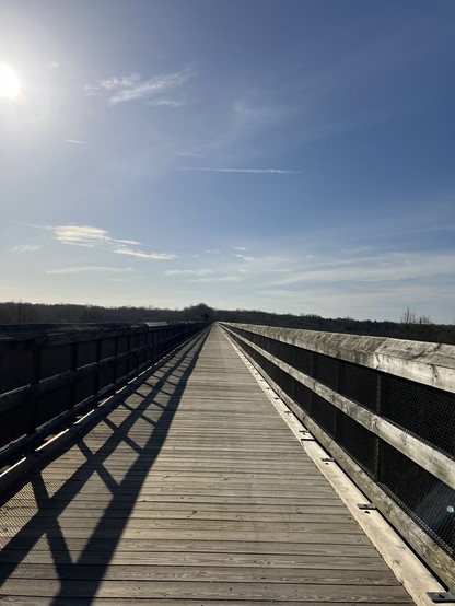 The railroad trestle bridge is now overlaid with wood planks. Here, it extends into the distance, with crisscrossing shadow patterns under a blue sky with scattered clouds.