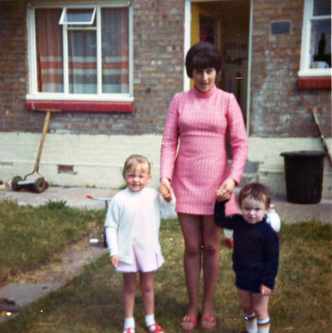 A scan of a 1970s colour photo in square format. The photo is taken on an overcast day in the back garden of a terraced house property. The garden has short but unkempt lawn with a narrow concrete path running down it. We're looking towards the back of the house which is mostly pale red brick except for the lower nine rows that have been painted white. There's an open yellow door up a couple of concrete steps and either side are single-glazed windows. The windows on the left have a small part open in the upper centre and we can see net curtains and curtains behind the glass. Under the window, against the wall, is a manual lawn mower with long handle and two wheels either side of the rotating blades. On the lawn in front of the house is a woman holding the hands of two children to pose for a photo. The woman has short dark hair and wears a pink, high-necked, tight-fitting dress that just reaches to her thighs. The child on the left is a young blonde girl wearing white and pale pink. The child on the right is a little smaller; a boy with blue shorts and jumper. He looks confused by what's occurring.