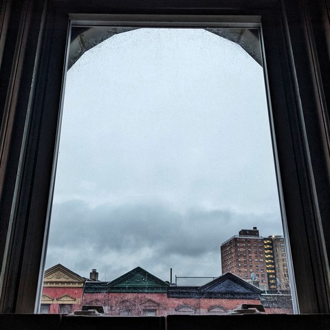 Looking through an arched window ten minutes after sunrise the flat grey sky has one thick band of grey cloud above the skyline. Pointed roofs of Harlem brownstones with red brickwork are across the street, and a taller apartment building can be seen in the distance. The window is grimy on the bottom.