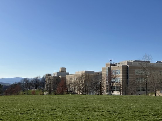 Large, modern buildings with a grassy lawn in the foreground and hills in the distance under a clear blue sky.