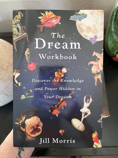 Photo of my new book titled "The Dream Workbook" by Jill Morris, featuring a night sky blue cover with a montage of surreal images like a fish with flames, floating objects, a moon, & a ballet dancer.