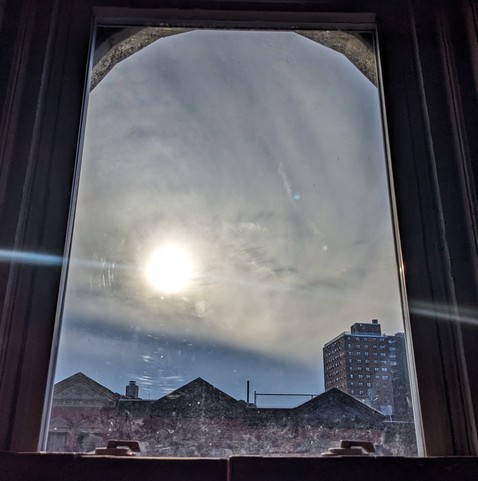 Looking through an arched window ninety minutes after sunrise the sun is a pale yellow disc shining through a murky blue-gray sky. Pointed roofs of Harlem brownstones with red brickwork are silhouetted across the street, and a taller apartment building can be seen in the distance. The window is grimy on the bottom.