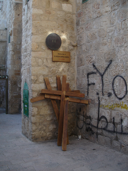 Fyra kors står uppställda lite slarvigt i ett hörn på en trång stadsgata. Ovanför korsen hänger en skylt som pekar ut riktningen mot St. Helen's coptic church. På väggen syns graffiti med latinska och arabiska bokstäver. Foto av Jon Brunberg  2009.