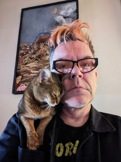 A middle-aged white man with an orange faux hawk and orange tinted glasses looks directly at the camera with a ruddy Abyssinian cat nuzzling on his shoulder. On the wall behind them is a depiction of the Fukushima disaster in Japan.