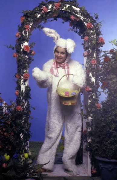 Bizarre pic of smiling Ozzy Osbourne dressed in a giant white rabbit costume with a pink satin bow at the throat. He's holding an Easter basket & he's standing under a garden arch wrapped in plastic roses. 