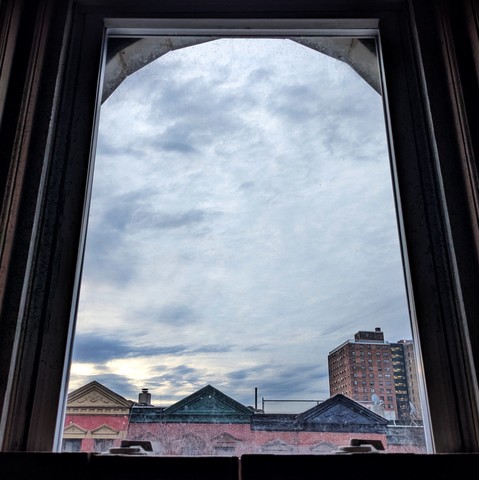 Looking through an arched window sixteen minutes after sunrise the sky is filled with sketchy scattered blue-gray clouds. Pointed roofs of Harlem brownstones with red brickwork are across the street, and a taller apartment building can be seen in the distance.