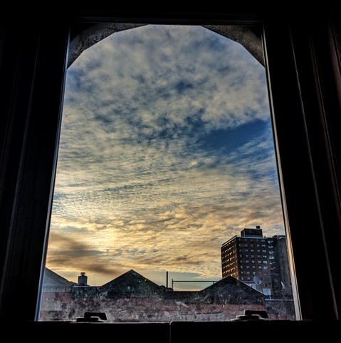 Looking through an arched window twenty- three minutes after sunrise small parchment colored clouds are sweeping across the blue sky. Pointed roofs of Harlem brownstones with red brickwork are silhouetted across the street, and a taller apartment building can be seen in the distance. The window is grimy on the bottom. 