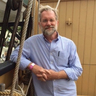 A man wearing glasses, a beard, and a button-down shirt leans against ship rigging.