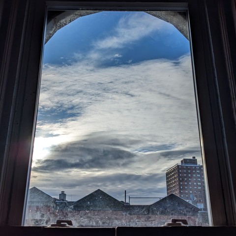 Looking through an arched window an hour after sunrise giant horizontal strokes of white and grey clouds fill most of the blue sky. Pointed roofs of Harlem brownstones are across the street, and a taller apartment building can be seen in the distance. The window is grimy on the bottom. 