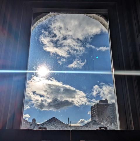 Looking through an arched window two hours after sunrise the blue sky is full of puffy white clouds and the sun is shining directly from the middle left of frame creating a dramatic horizontal lens flare. Pointed roofs of Harlem brownstones are silhouetted across the street, and a taller apartment building can be seen in the distance. The window is grimy on the bottom. 