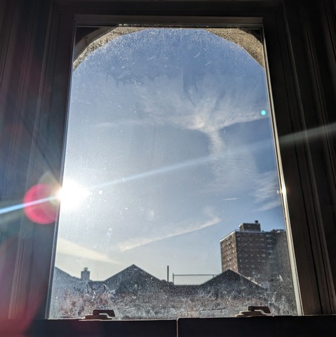 Looking through an arched window ninety minutes after sunrise the blue sky is marked with stringy white clouds and the sun is shining directly from the middle left of frame creating a dramatic horizontal lens flare. Pointed roofs of Harlem brownstones with red brickwork are silhouetted across the street, and a taller apartment building can be seen in the distance. The window is grimy on the bottom. 