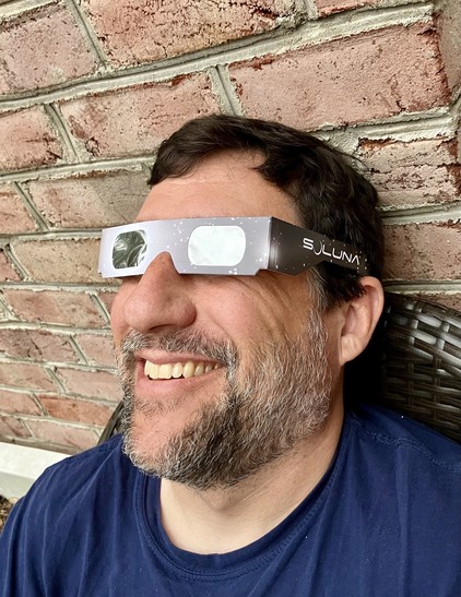 A smiling person wearing eclipse viewing glasses with a brick wall in the background.